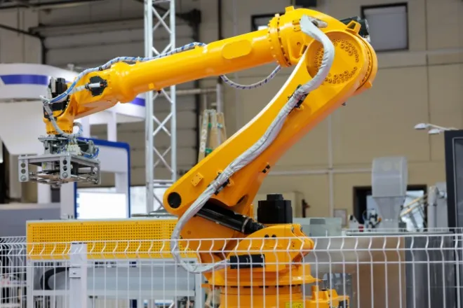 A yellow robotic arm of an automatic robotic palletizer system in a short cage inside of a warehouse.