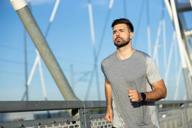A man in a sweat stained shirt is jogging in an outdoor environment. Behind him is a suspension system for a bridge.
