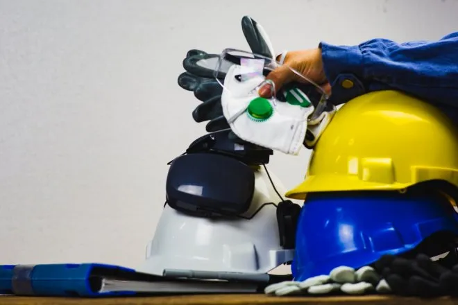 A stack of PPE equipment resting on a table. Someone holds a face mask, safety glasses, and gloves on top of some hard hats.