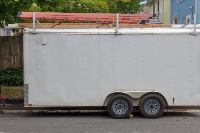 A white work trailer parked in a neighborhood on the curb. There is an orange ladder secured to the roof rack.