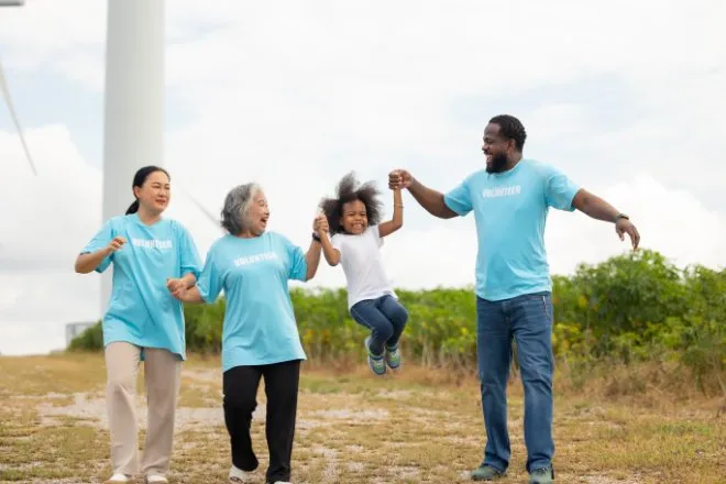 Three volunteers in blue shirts swing a laughing child by the hands on a path near a rural wind turbine and field.