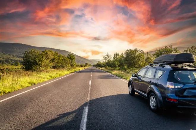 A car is driving down a road. On either side of the road are rows of trees and there are rose-colored clouds in the sky.