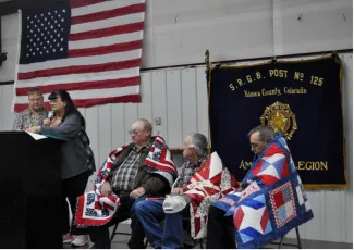 Military veterans seated on a stage wrapped in quilts listening to presenters.