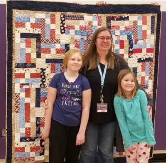 Amanda Plitz with daughters Isabelle and Chloe. Courtesy Rayetta Addy Woman and two children standing in front of a quilt.