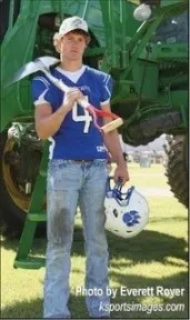 Young man wearing a sports jersey holding a shovel and football helmet in front of a piece of large farm equipment.