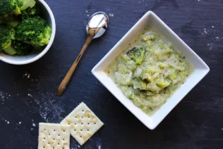 Cream of Broccoli Soup. Square bowl of Cream of Broccoli Soup, a bowl of broccoli, two crackers and a spoon.