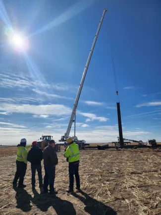 Group of construction workers and a crane setting a power pole in a field.