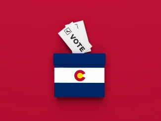 PROMO HIRES Election Colorado Vote Ballot - Baris-Ozer - iStock-1420488382 Miniature ballot box with and image of the Colorado state flag on a red background with slips of paper representing ballots
