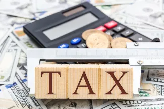 PROMO HIRES Government - Taxes Money Calculator Word Blocks - iStock - LIgorko Word "tax" on wooden blocks held together with a caliper tool. A calculator and assorted coins and currency are in the background.
