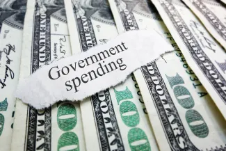 PROMO HIRES Finance - Government Spending Words Budget Money Cash - zimmytws - iStock-511889079 A scrap of torn paper with the words "Government spending" rests on top of a spread of United States 100 dollar currency.