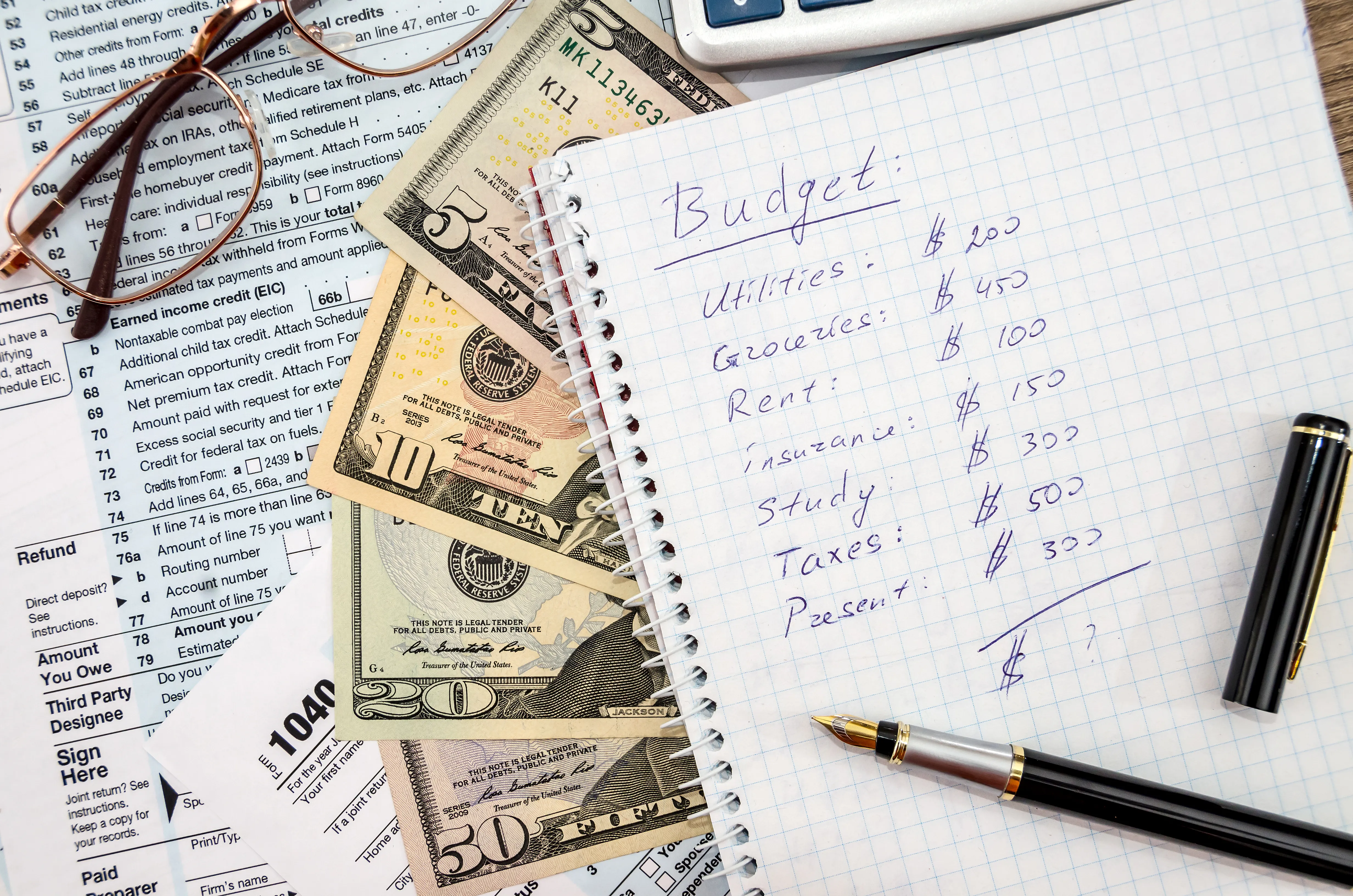 Notebook with a pen on a page showing a personal budget. Underneath are paper currency and tax forms. Eyeglasses are nearby