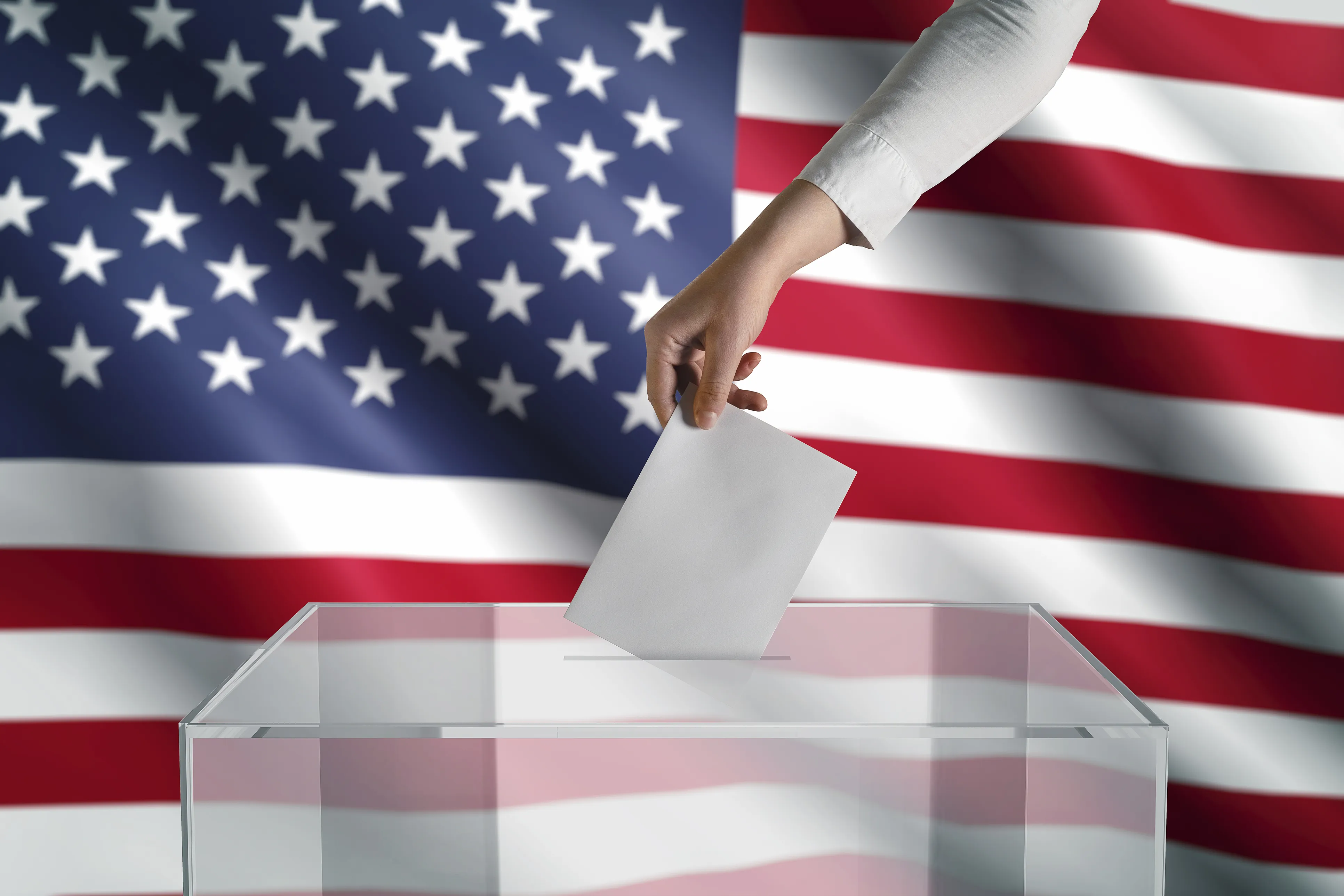 Hand placing a piece of pager into a clear ballot box in front of the United States flag