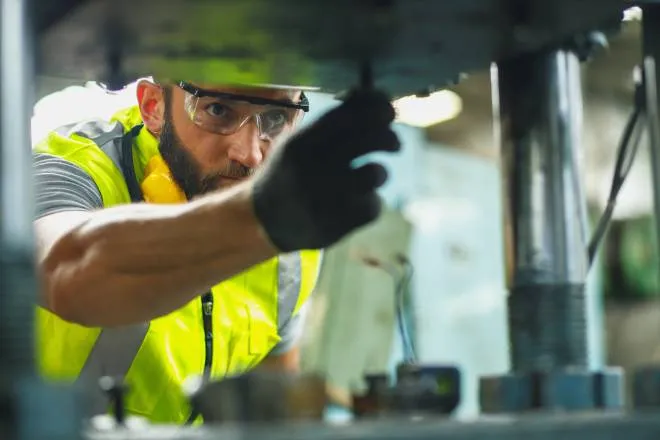 A man in high-visibility gear, safety glasses, and gloves turning hardware on a piece of industrial equipment.
