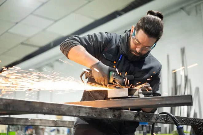 A man working hard in a machine shop with a glass grinder machine and sparks flying. He's wearing proper PPE.