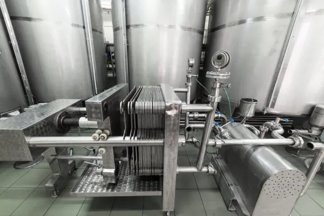 A large stainless steel automatic filter press sits in a room with large stainless steel storage containers.