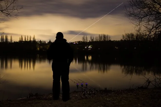 An angler standing at the edge of a lake just after sunset with three fishing rods resting on the ground next to him.