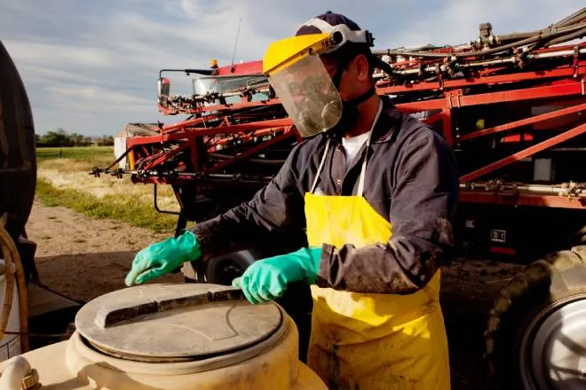 A person wearing a protective shield, a yellow protective apron, green protective gloves, and a mask is opening a container.