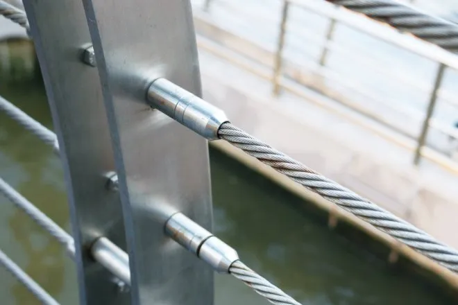 A close-up of a stainless steel threaded stud joint with wire rope cable railing over top of a body of water.