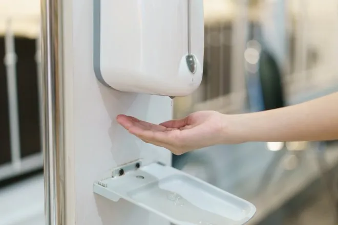 A touchless soap dispenser attached to a bathroom wall next to a mirror. A cupped hand is reaching out underneath it.