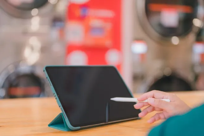 Someone using a stylus on their propped-up tablet at a laundromat. There are laundry machines blurred out in the background.