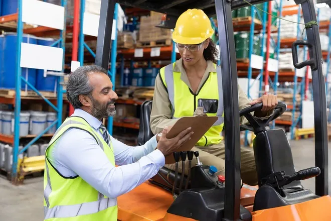 Two workers wearing safety vests. One is sitting in a forklift while the other is showing them information on a clipboard.