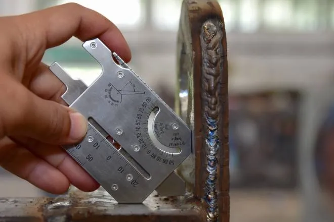 A technician uses a welding gauge to measure the dimensions of a metal part. The metallic joint lines are visible.