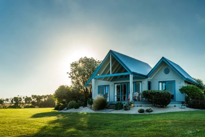 A luxurious home sits on a wide and empty plot of land. There is greenery around the home and the sun behind it.