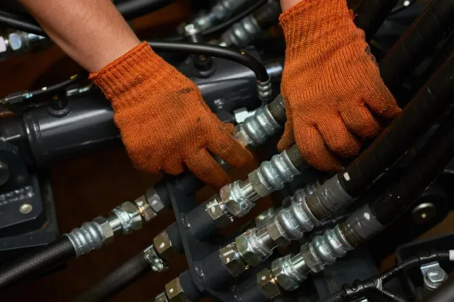 Close-up of a person's hands maintaining a hydraulic hose system. The person wears dirty orange gloves on their hands.