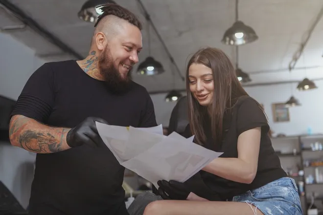 A tattoo artist wearing black latex gloves showing a smiling client several printed images for them to consider.