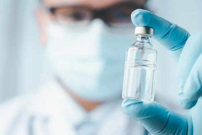 A scientist wearing a blue latex glove holding a small glass sample vial filled with a clear liquid in front of his face.