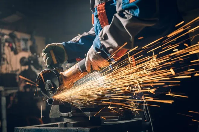A metalworker using a saw to cut something. The worker is in safety gear and there are sparks flying back at them.