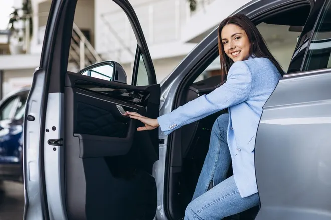 A woman in a blue suit jacket sits in the front seat of a silver car with the door open.