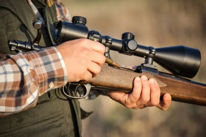  A close-up of a hunter, wearing a flannel shirt and vest, loading a round into a bolt-action carbine with a scope.