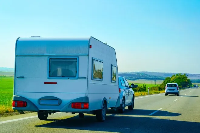  A white car towing a camper on a highway. There is an empty green field around the road and a clear blue sky overhead.