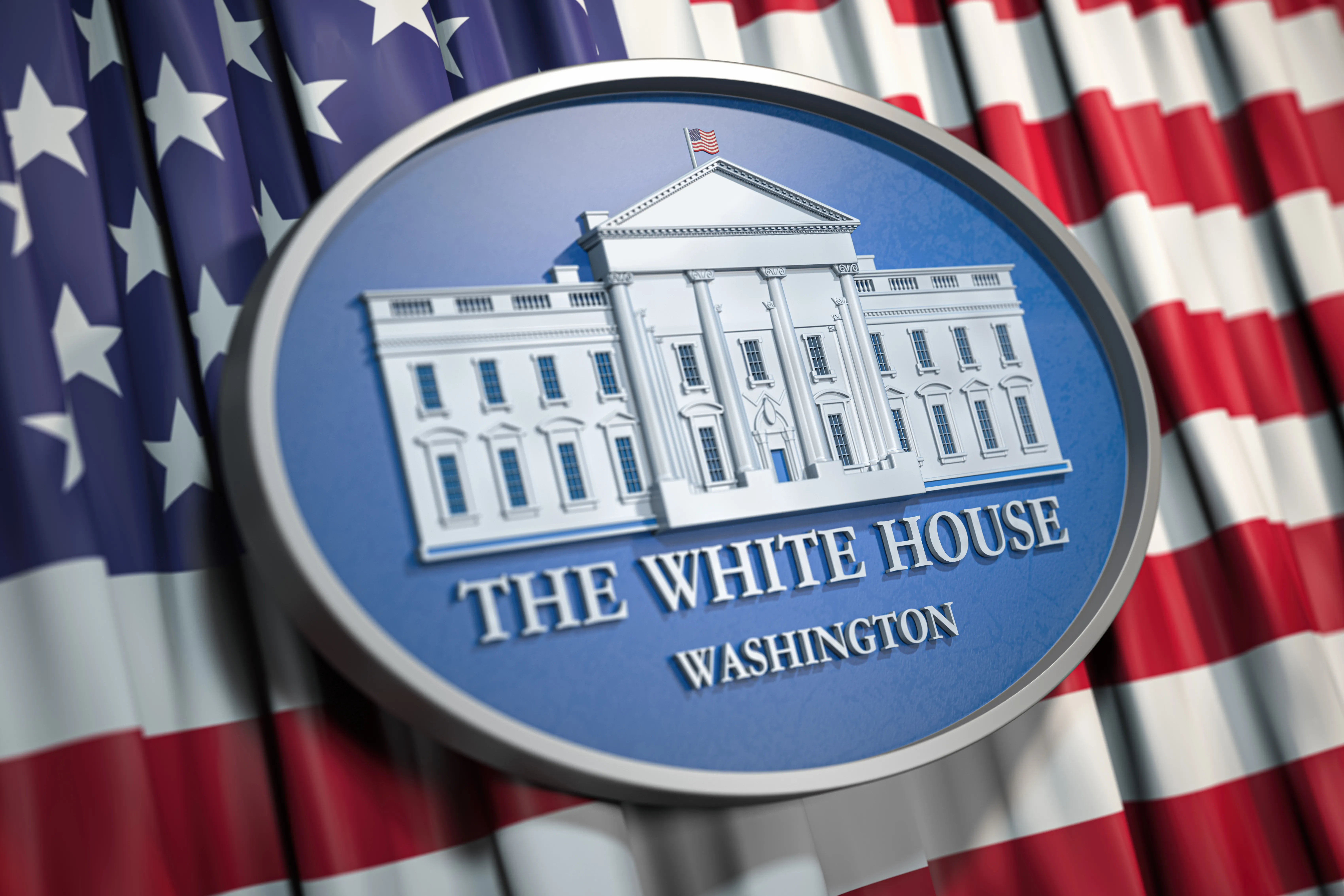 Close up of an oval, carved plaque with an image of the White House. The plaque is on top of a flag of the United States of America.