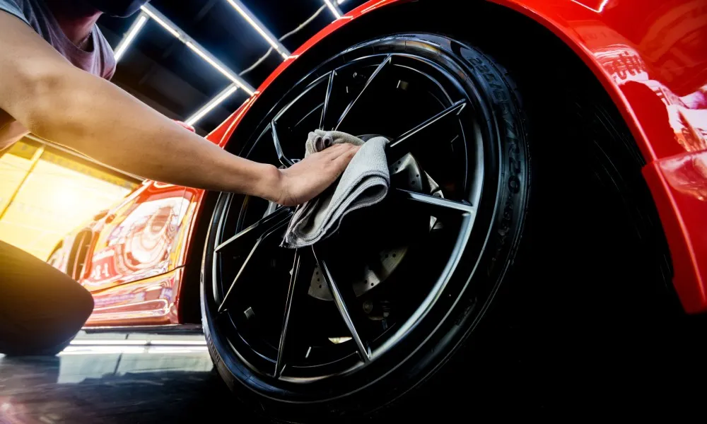 A man is using a washcloth to clean the spokes of a car tire. The car is red and the man’s face is obscured by a mask.