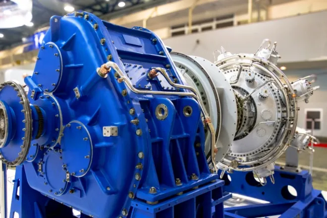 A close-up of a large blue industrial gearbox connected to a turbine unit inside a factory with overhead lighting.