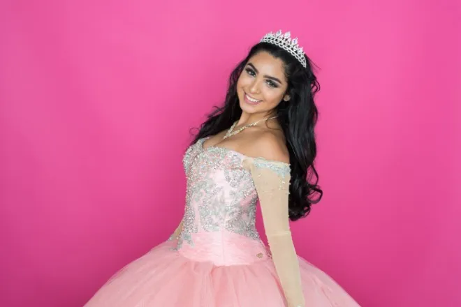 A girl with long, dark hair wearing a tiara and a formal pink dress poses against a bright pink background.