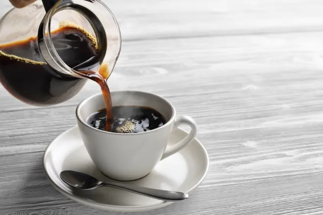 A close-up of a glass coffee pot pouring hot coffee into a white mug on a saucer. A silver spoon rests on the saucer.