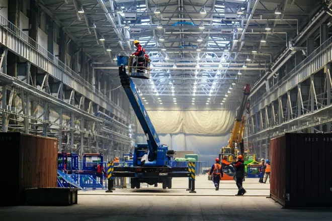 Several people are working in a large industrial space with visible equipment and material around them.