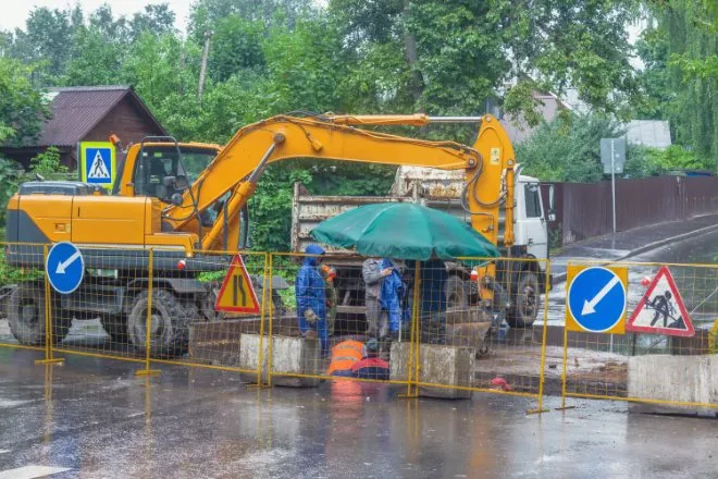 An excavator and dump truck parked behind a yellow fence with different signs. Crew members wear blue ponchos as it rains.