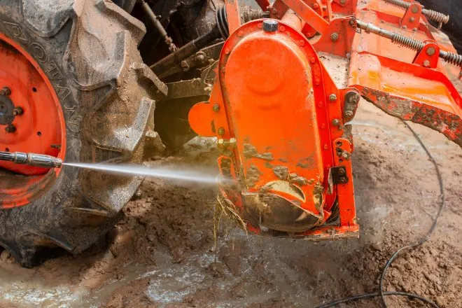 A pressure washer sprays the surface of a muddy red tractor attachment beside a tire. Water pools on the ground.