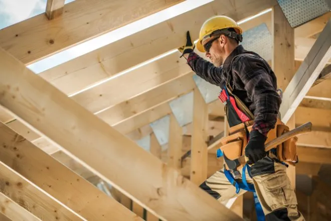 A man in a hardhat is standing n the rafters of a structure. He’s looking at something off-screen.