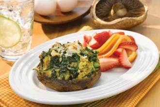 Herbed Spinach Quiche Portabella Caps - USDA Plate on a placemat with a herbed spinach quiche Portabella mushroom cap and sliced strawberries and oranges, surrounded by a glass of water, small wooden plate with two eggs, and an uncooked Portabella cap.