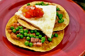 Plate with two corn tortillas with refried beans, ham, and peas, topped with a slice of cooked eggs and salsa.