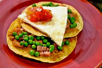 Huevos Motulenos - USDA Plate with two corn tortillas with refried beans, ham, and peas, topped with a slice of cooked eggs and salsa.