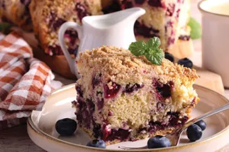 Slice of blueberry coffee cake on a plate with napkin and small cream pitcher in the background.