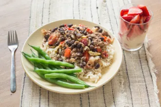 Picadillo Plate on a placemat of picadillo on a bed of rice with green beans. A fork and glass of sliced strawberries are next to the plate.