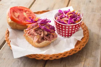 Wicker basket lined with a paper towel. A container of red cabbage and carrot slaw sits to one side, accompanied by a bread bun with pulled pork on one half and a tomato slice on the other half.