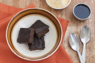 Sweet Cornmeal Pudding with Ginger and Cinnamon - USDA Overhead view of a bowl on a table with three squares of sweet cornmeal pudding. Spoons and small bowls of condiments are nearby.